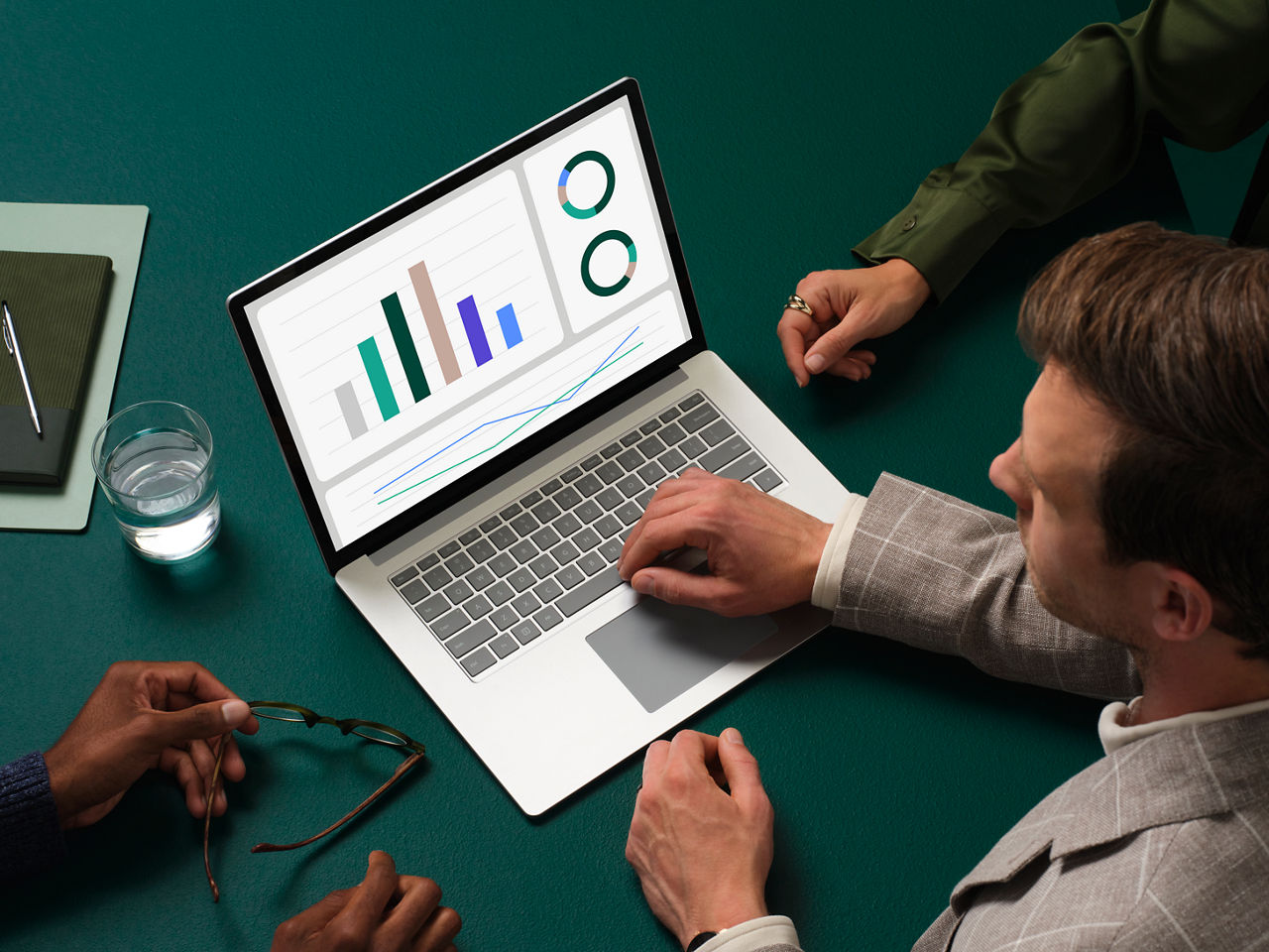 A laptop computer with colorful graphs and charts on screen sits on a dark green conference table where one man is controlling the laptop and two other people sit next to him.