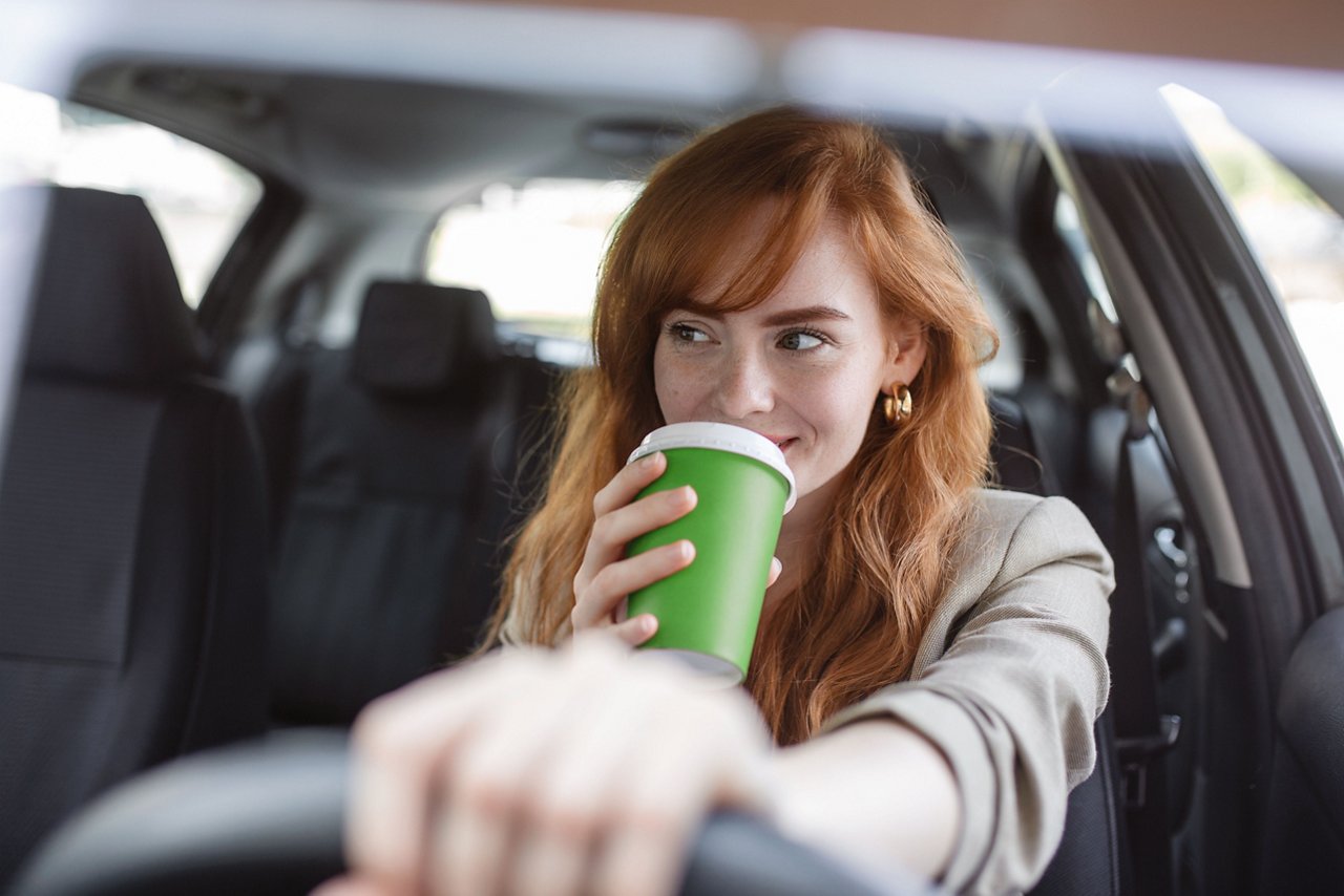 Happy young woman with takeaway coffee driving her car. Woman sipping coffee while driving a car. Young woman drinking coffee while driving her car. Attractive redhead driving a car; Shutterstock-ID-2155032721; purchase_order:Kreditkarte; Job:Tim Hortons Karton Studie FUZE Page und Ellen McPartlan; client:pgarvey@spotcreates.com; andere:USA