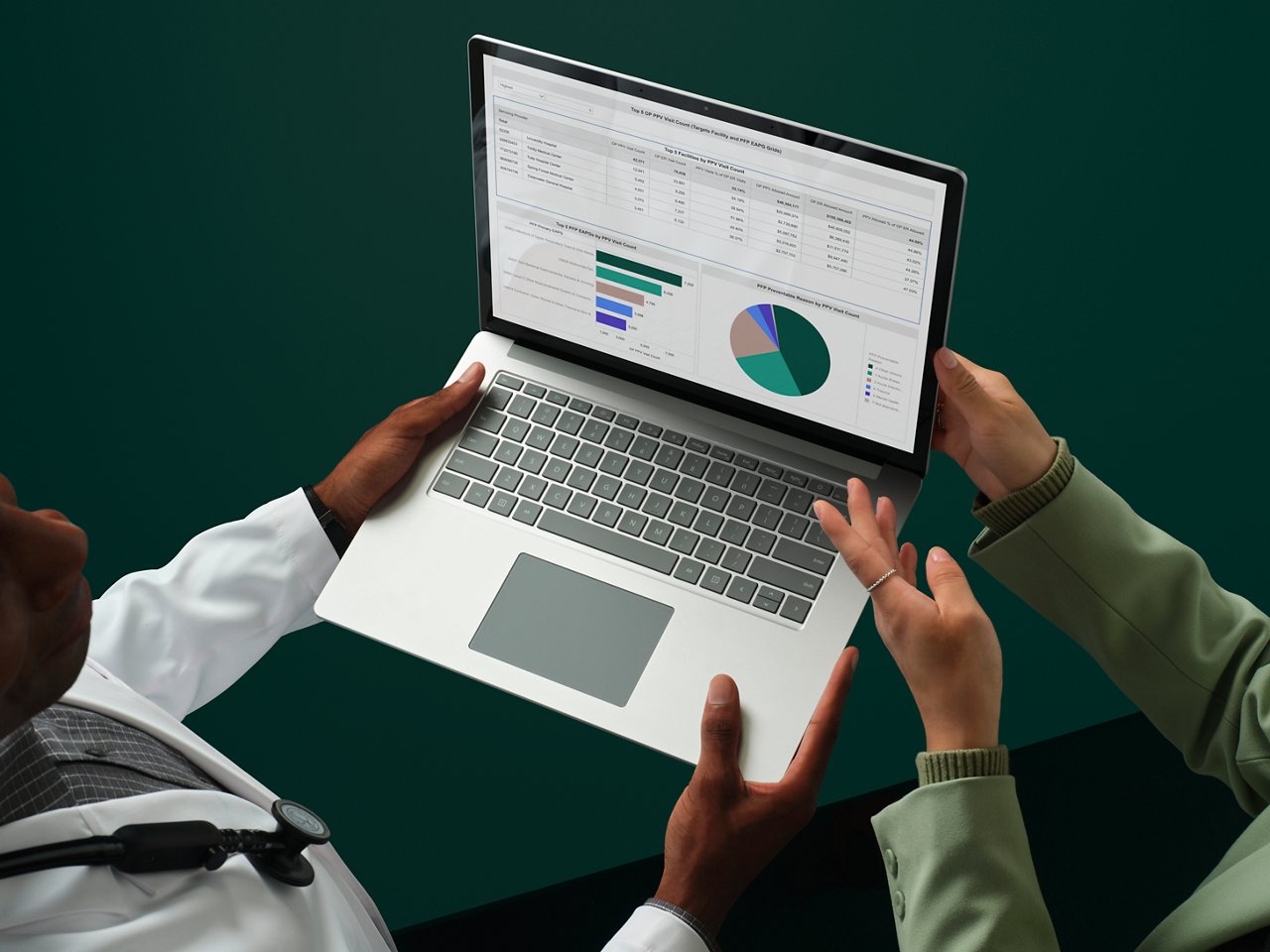 At a dark green conference table, a male physician in a white lab coat holds a laptop computer with colorful graphs and charts on screen while a woman in business clothes gestures toward the screen.