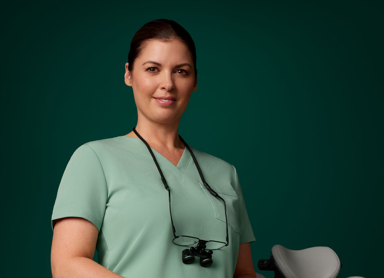 Portrait of an EMEA female dentist in scrubs standing next to a dental chair on a dark green background.
