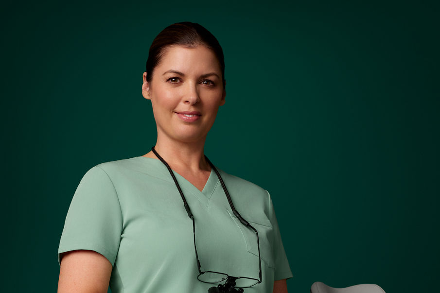 A woman in a lab coat with glasses and a loupe on a leash around her neck sits and smiles.
