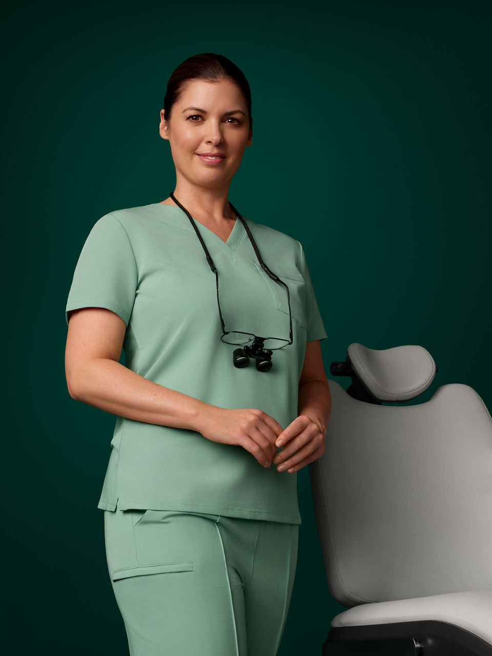 Portrait of an EMEA female dentist in scrubs standing next to a dental chair on a dark green background.