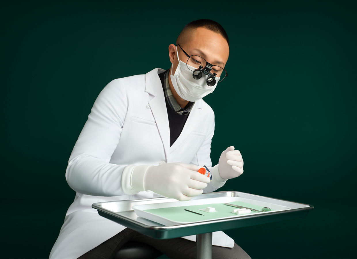 Male dentist sitting behind dental tray on stand using Scotchbond™ Universal Plus Adhesive, dark green background.
