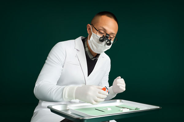 Male dentist sitting behind dental tray on stand using Scotchbond™ Universal Plus Adhesive, dark green background.