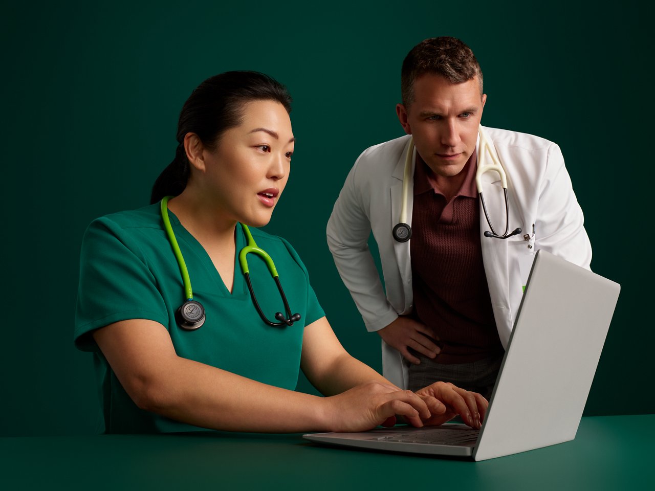Portrait of a male doctor and female nurse viewing a laptop screen on a dark green background.