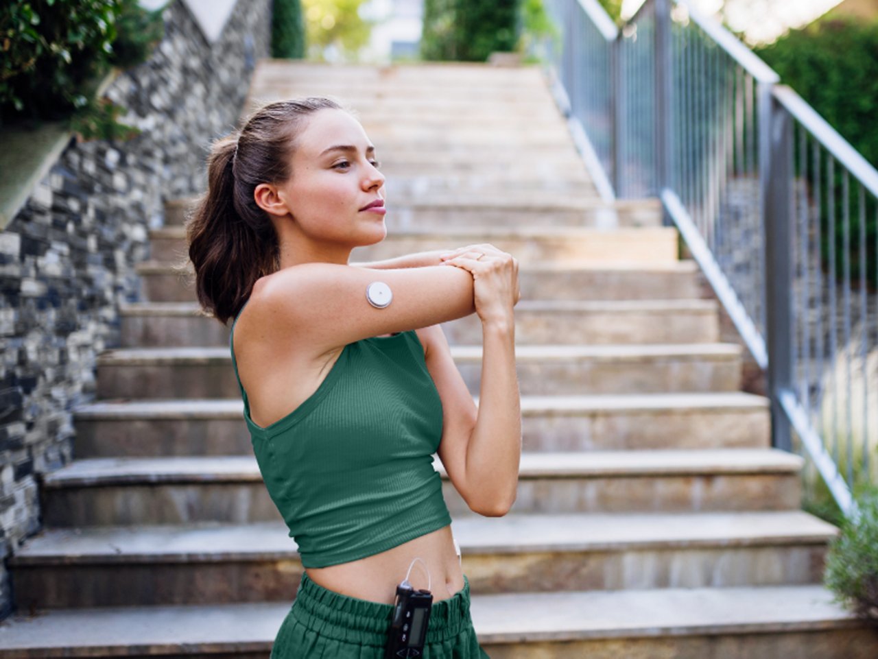 一位女士在楼梯附近伸展双臂，右臂上佩戴着小号圆形的监控葡萄糖仪。
