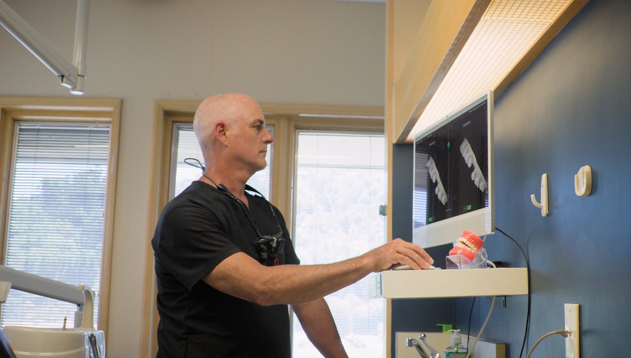 Dr. Marc Geissberger at his dental clinic wearing black scrubs and dental loupes, reviewing 3D dental scans on a computer.