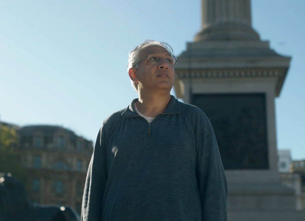 A man stands in front of a monument with a column and statue on a clear, sunny day.