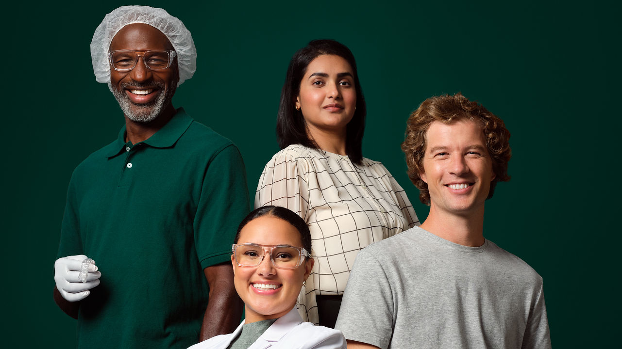 Photo of a enterprise employees. A female application engineer wearing eye glasses and blue gloves seated, a male HISD employee seated on a stool and holding a software device, a male OCSD manufacturing employee in a hair net and holding an aligner case standing alongside an MSD female employee.