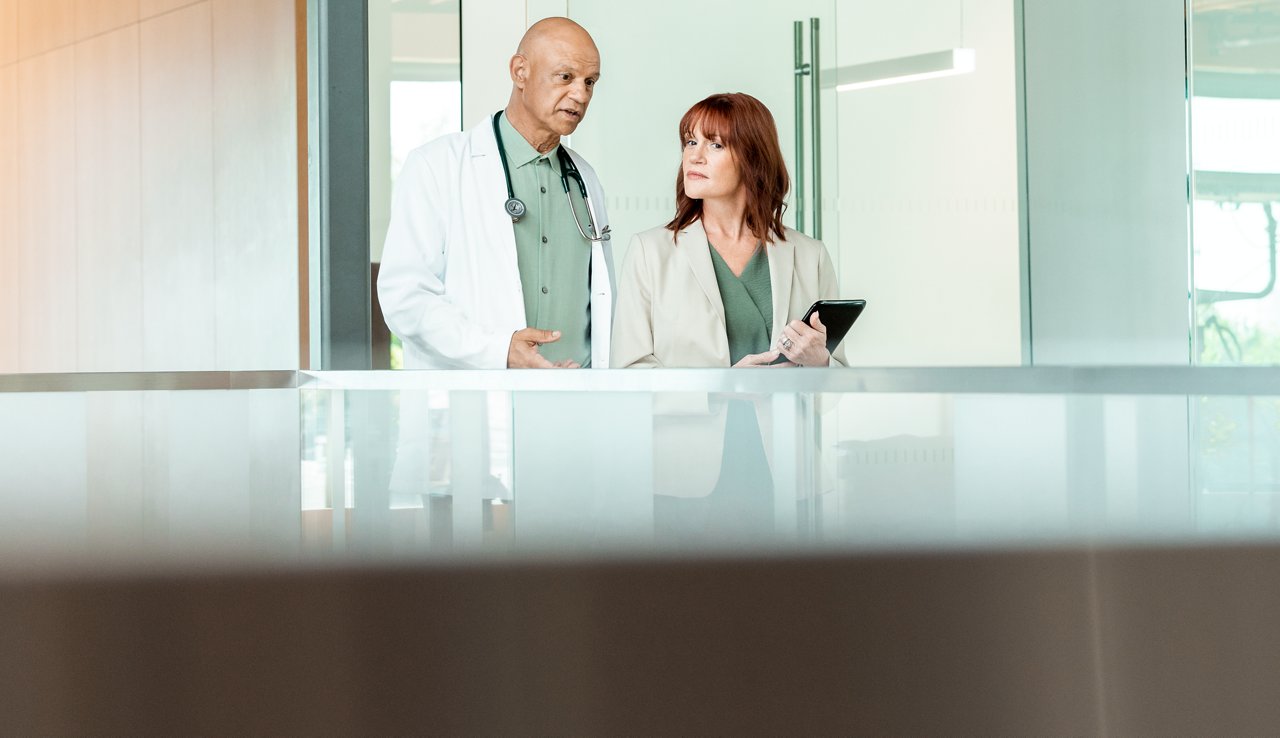A male doctor, wearing a lab coat and Littman stethescope, walks alongside and talks to a female admin holding a tablet.
