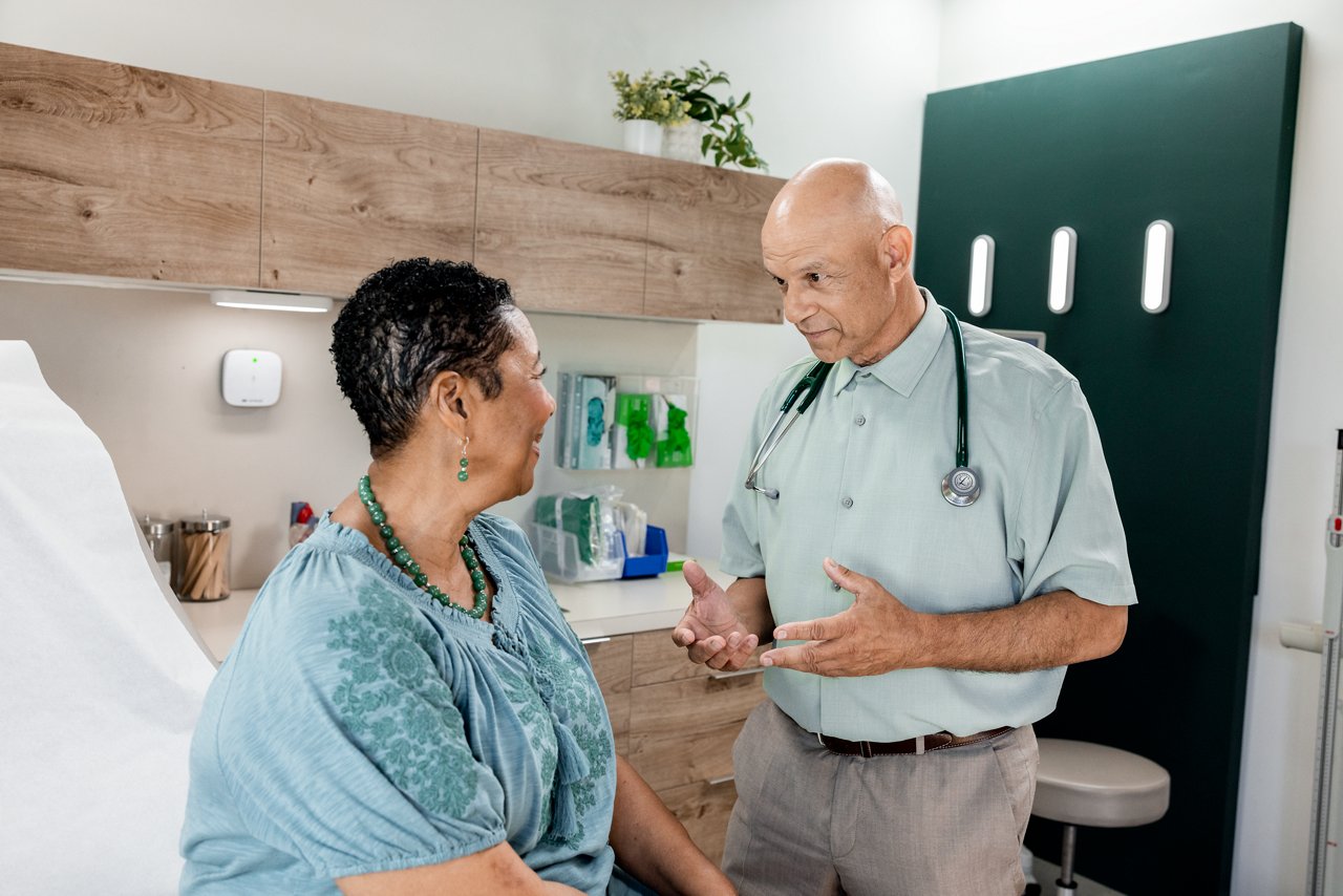 Mature female patient interacting with a male primary care doctor not wearing a jacket in an exam room.