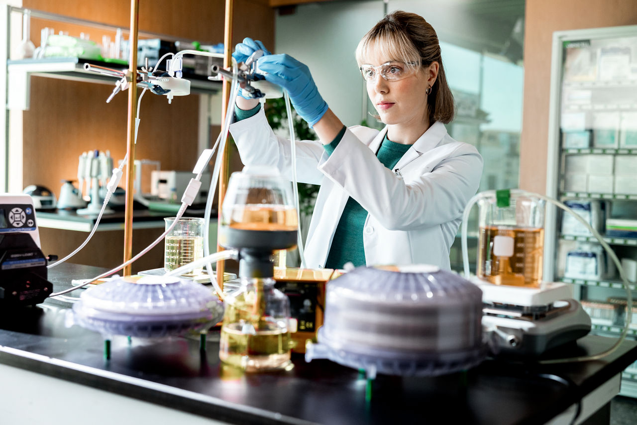 A female scientist wearing a lab coat, safety glasses and gloves works at a lab station.