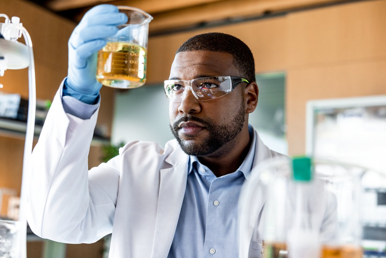 A male scientist wearing a lab coat, safety glasses and gloves holds a beaker at eye-level to examine the contents.