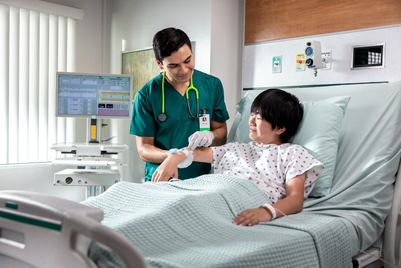 A male nurse wearing a Littman stethoscope checks the dressings on a young male patient in a hospital gown and bed.