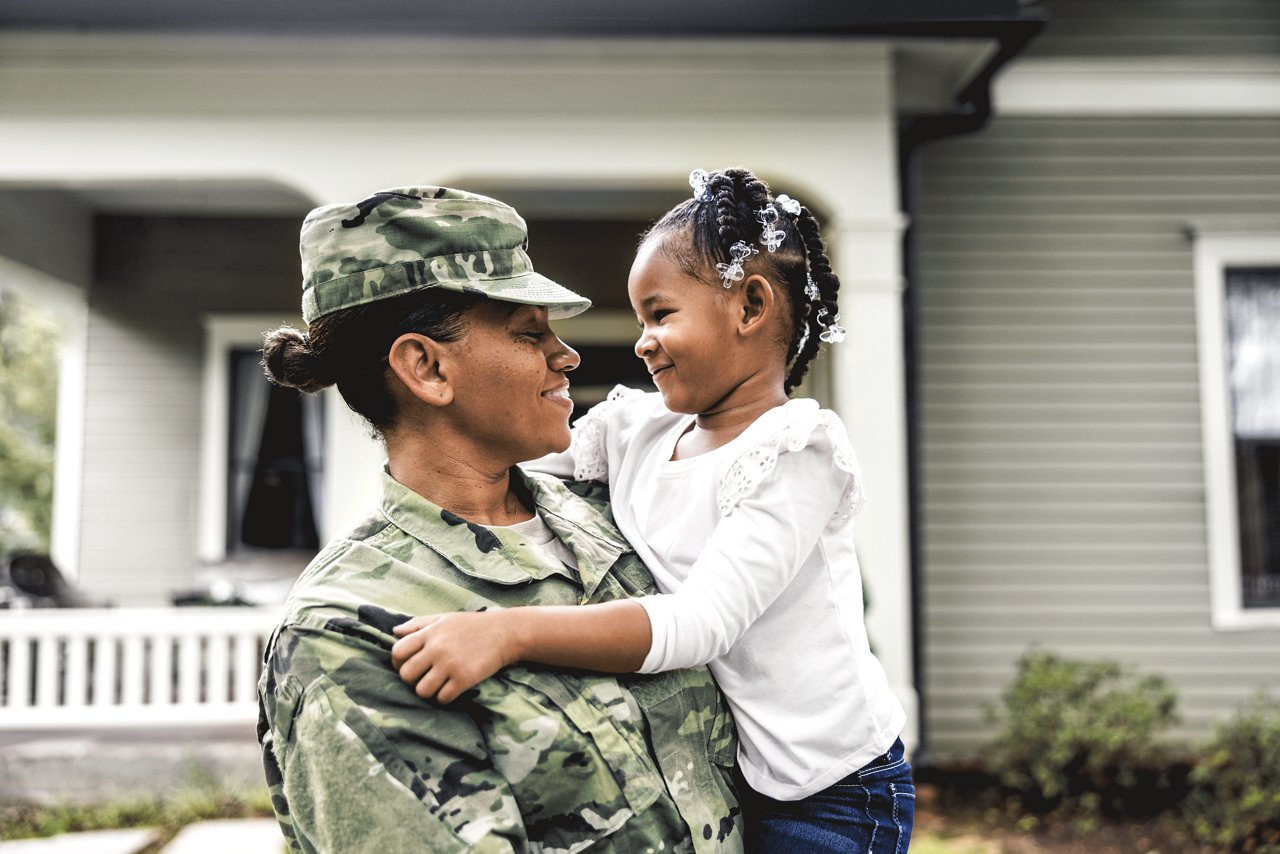 A woman in camouflage military fatigues holds her young daughter in her arms.