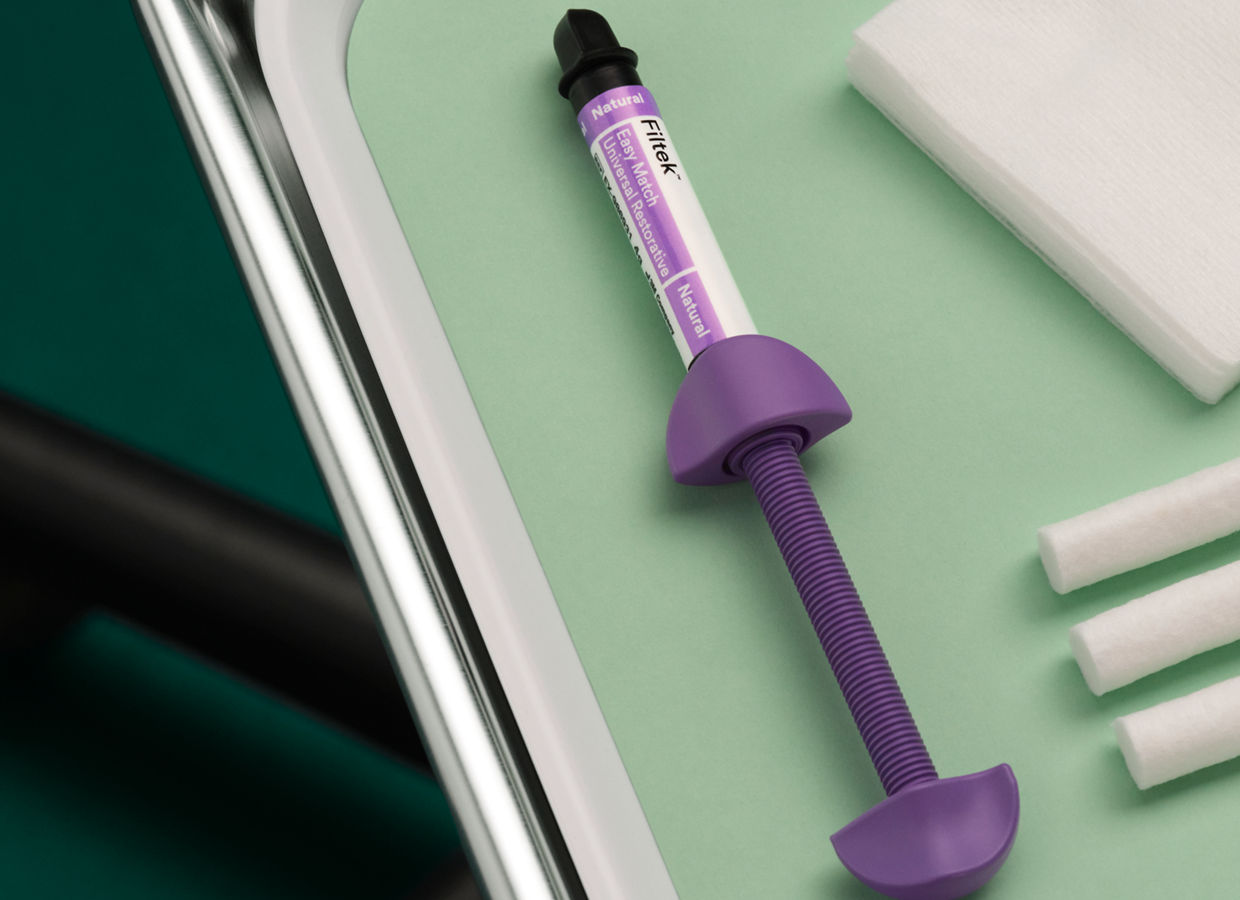 3M Filtek Easy Match Universal Restorative syringe lays on a metal dentist tray with white plastic insert and light green liner, floor background is dark green.