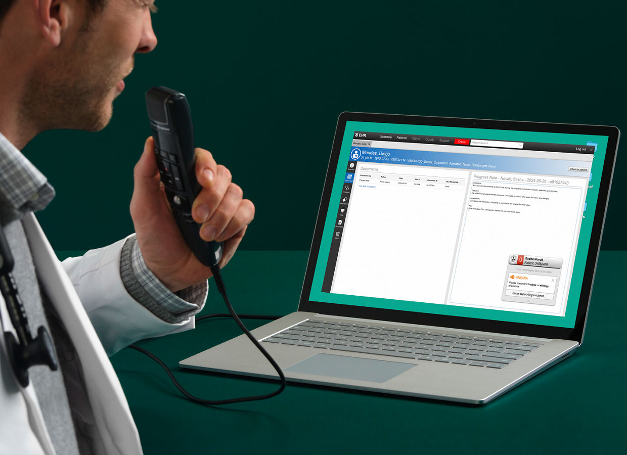 Un médico está sentado en una mesa verde oscuro mientras habla en un dispositivo de dictado de Solventum que está conectado a un ordenador portátil con una aplicación de software en la pantalla.