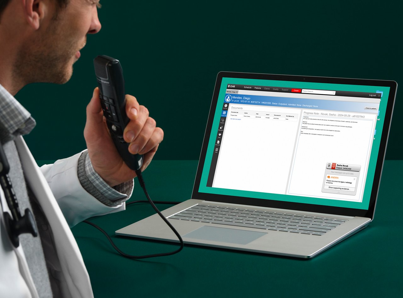 Male physician sits at a dark green table while speaking into a 3M M*Modal Fluency Direct dictation device that is hooked up to a laptop computer with software application on screen.