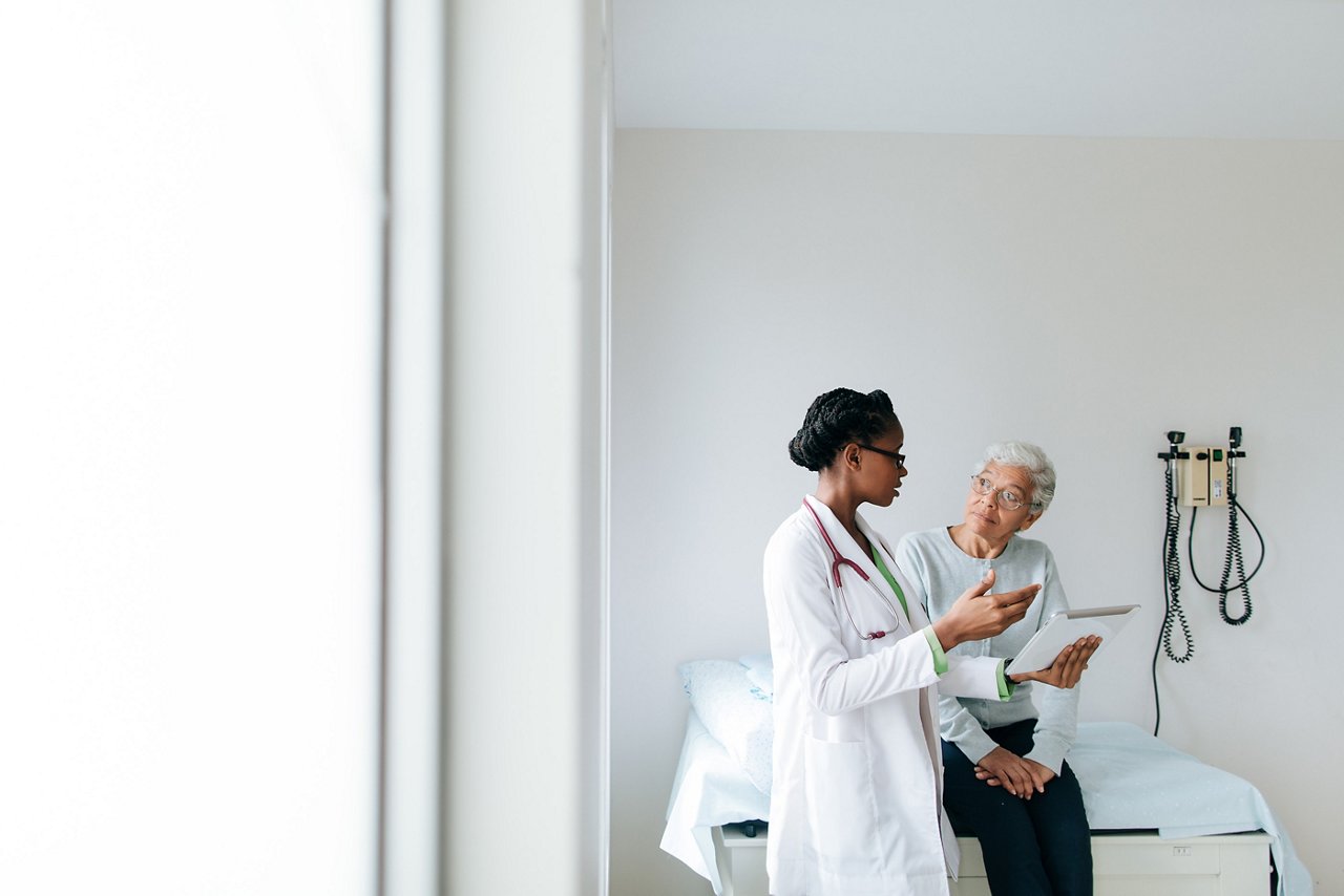 Ein Kliniker steht neben einem älteren Patienten auf einer Untersuchungstabelle und erklärt Informationen auf einer Tablett-Maske.