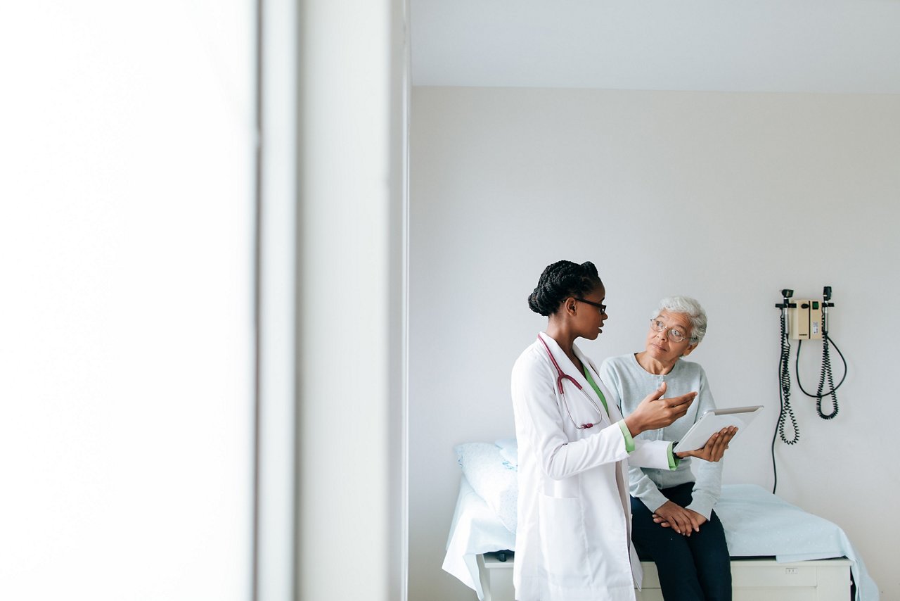 A clinician standing beside an older patient on an exam room table, explaining information on a tablet screen.