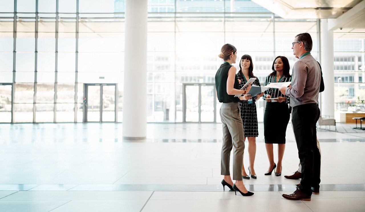 Group of five colleagues chatting while standing in an office lobby.