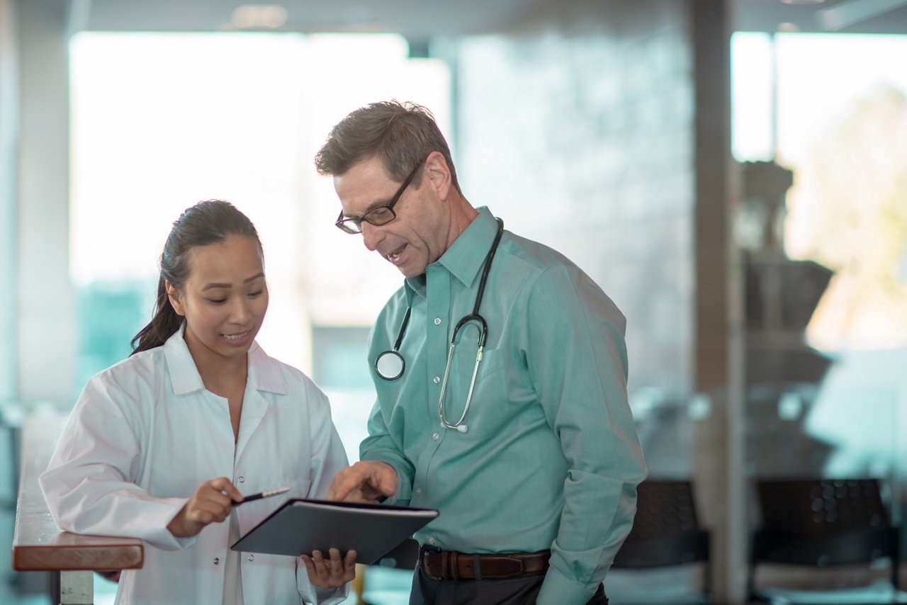 A woman in a white medical coat and a man in business clothes with a stethoscope around his neck looking at a tablet