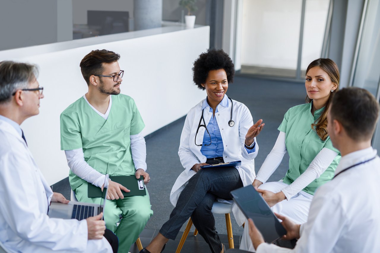 A group of healthcare professionals including doctors, nurses and specialists seated in a circle who are interacting. A couple of them are smiling.