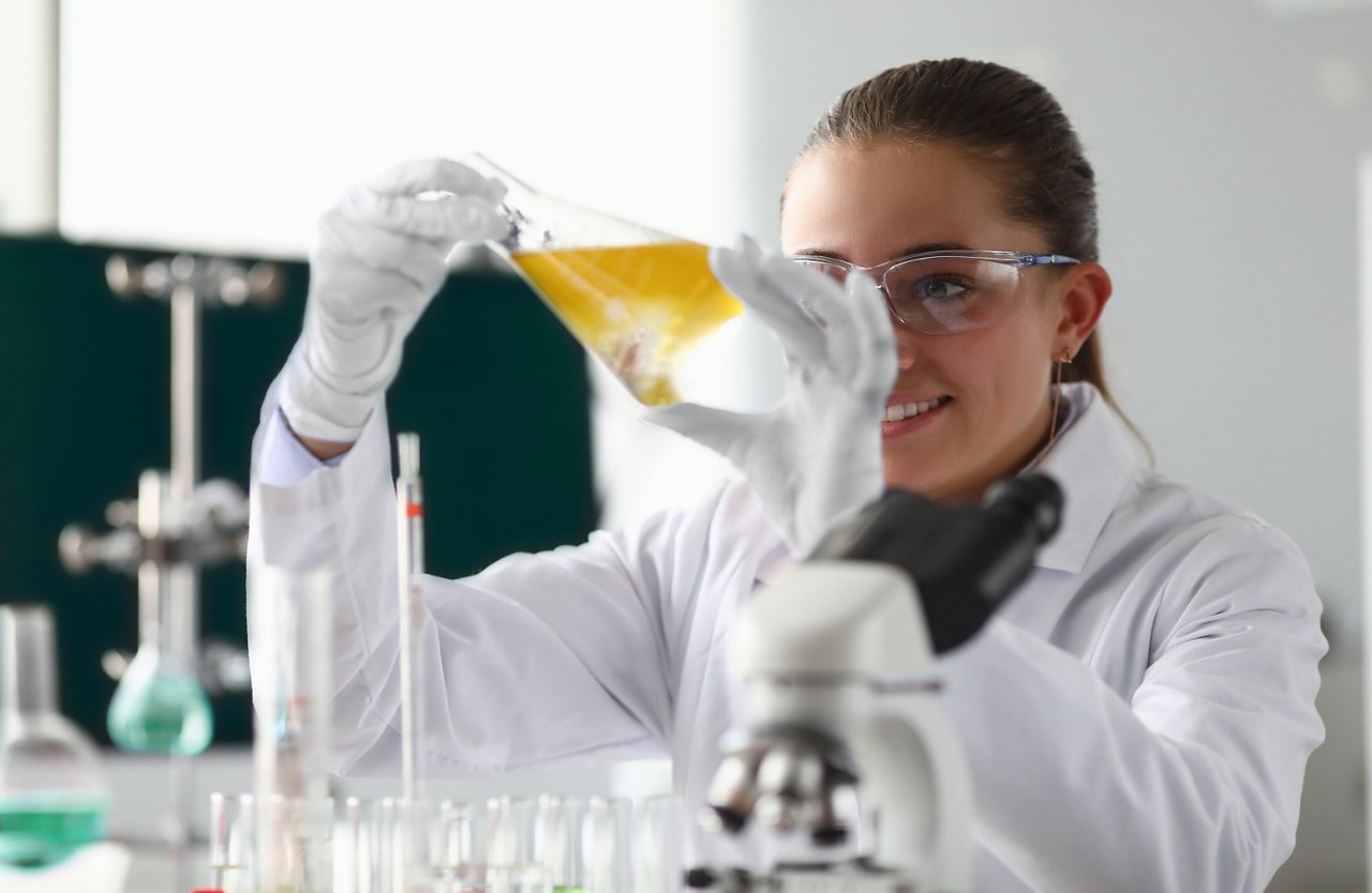 A scientist wearing safety goggles, gloves and a lab coat inspecting a beaker of liquid in a lab.