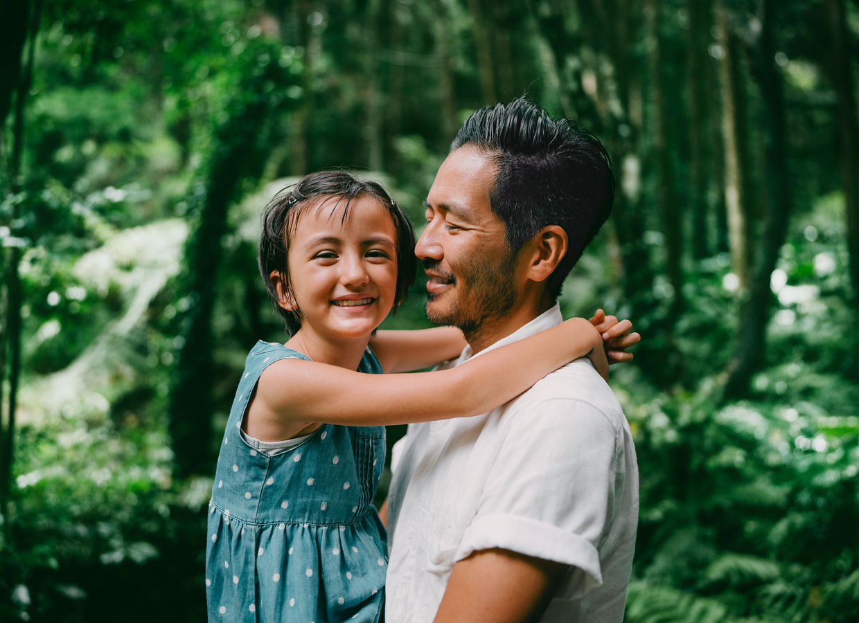 Padre sosteniendo a su hija pequeña en sus brazos rodeados de un bosque exuberante.