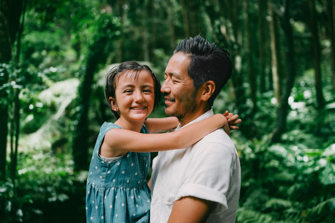 Padre sosteniendo a su hija pequeña en sus brazos rodeados de un bosque exuberante.