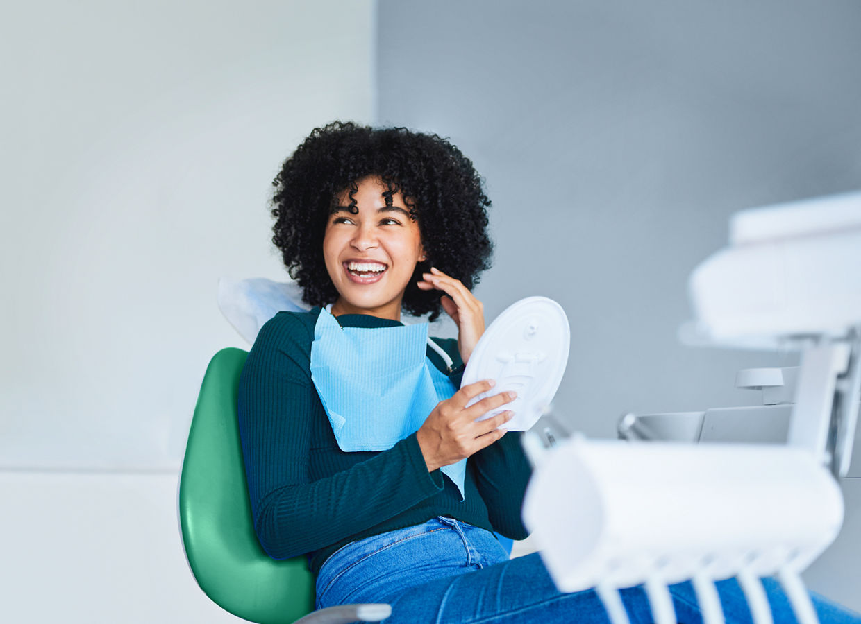 Uma jovem sorrindo sentada em uma cadeira odontológica com um espelho na mão.