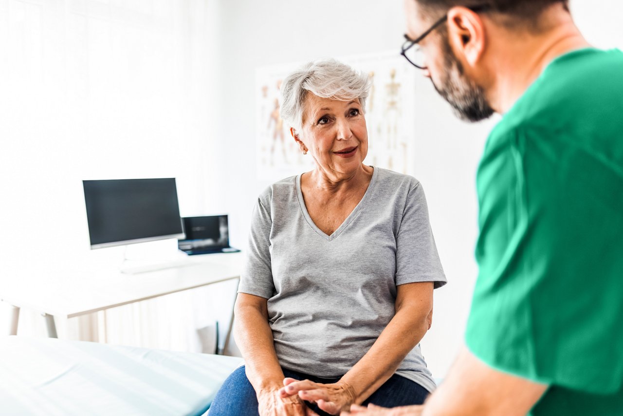 A doctor wearing scrubs consulting with an older patient who is seated on an exam table.
