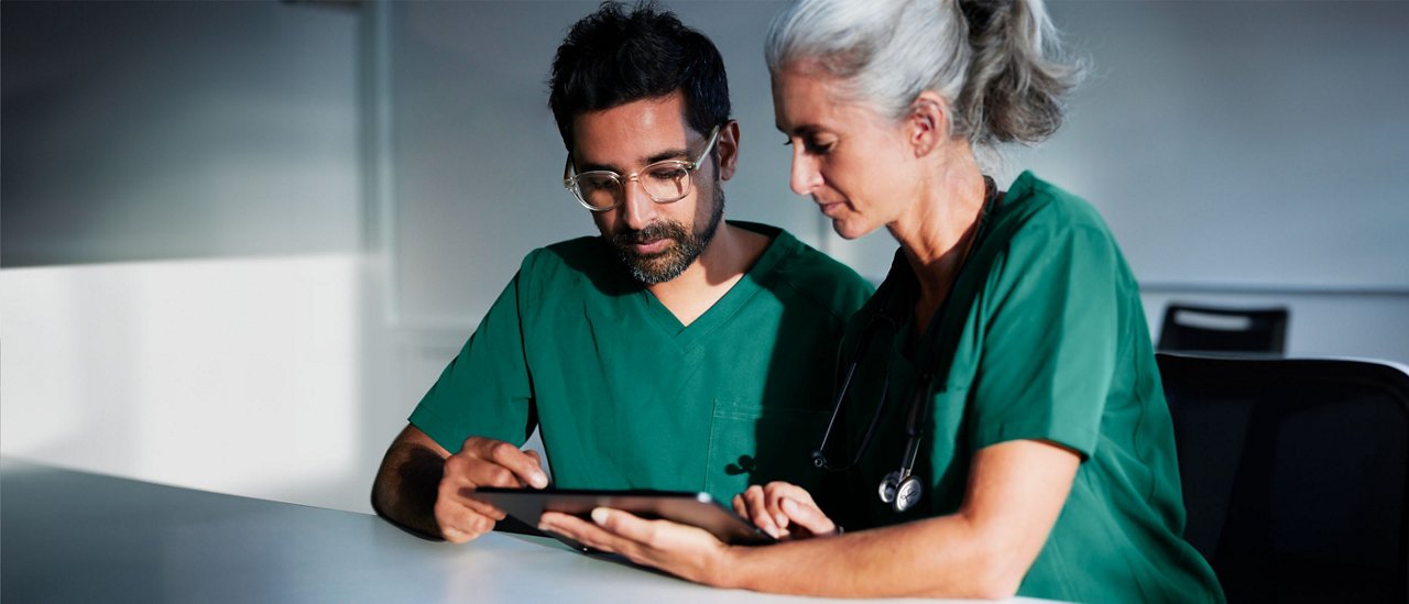 This is a Getty image that has been retouched to reflect the Solventum brand. It is an image of a female and male clinician both in scrubs viewing a digital tablet screen in a classroom setting.
