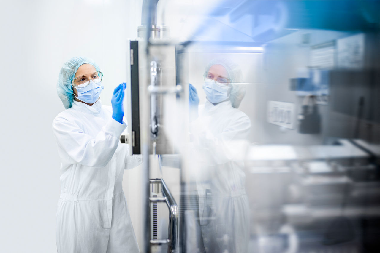 A sterile-gowned female worker in a clean room standing next to equipment and touching the screen.