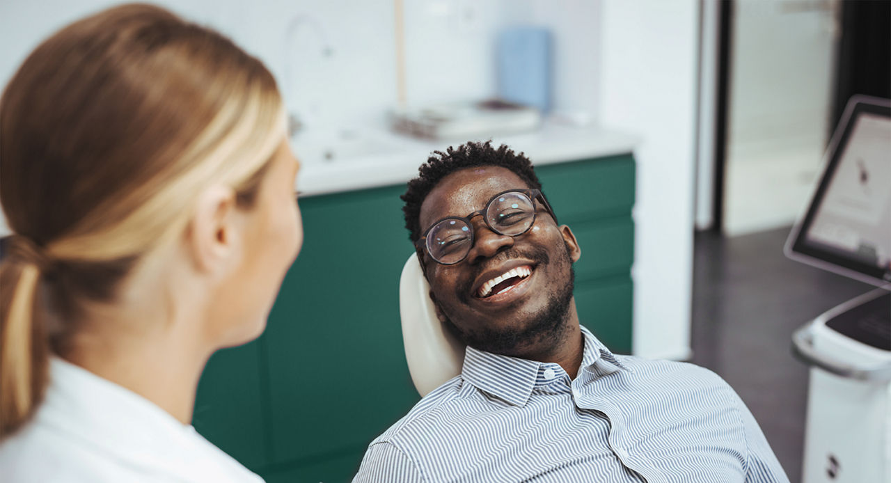Smiling african guy in dentist chair looking with trust at his doctor, close up. Young African American Man Getting Teeth Treatment With Professional Stomatologist At Modern Clinic