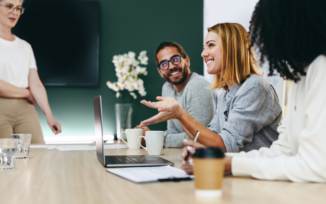 Varios colegas sonrientes conversando en una sala de conferencias. Uno de ellos está de pie y los demás están sentados en una mesa de conferencias.