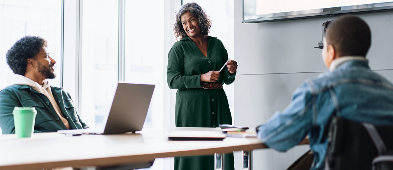 A smiling female presenter is standing at the front of a meeting room near the screen and a smiling male employee and another employee in a wheelchair are seated at a conference table.