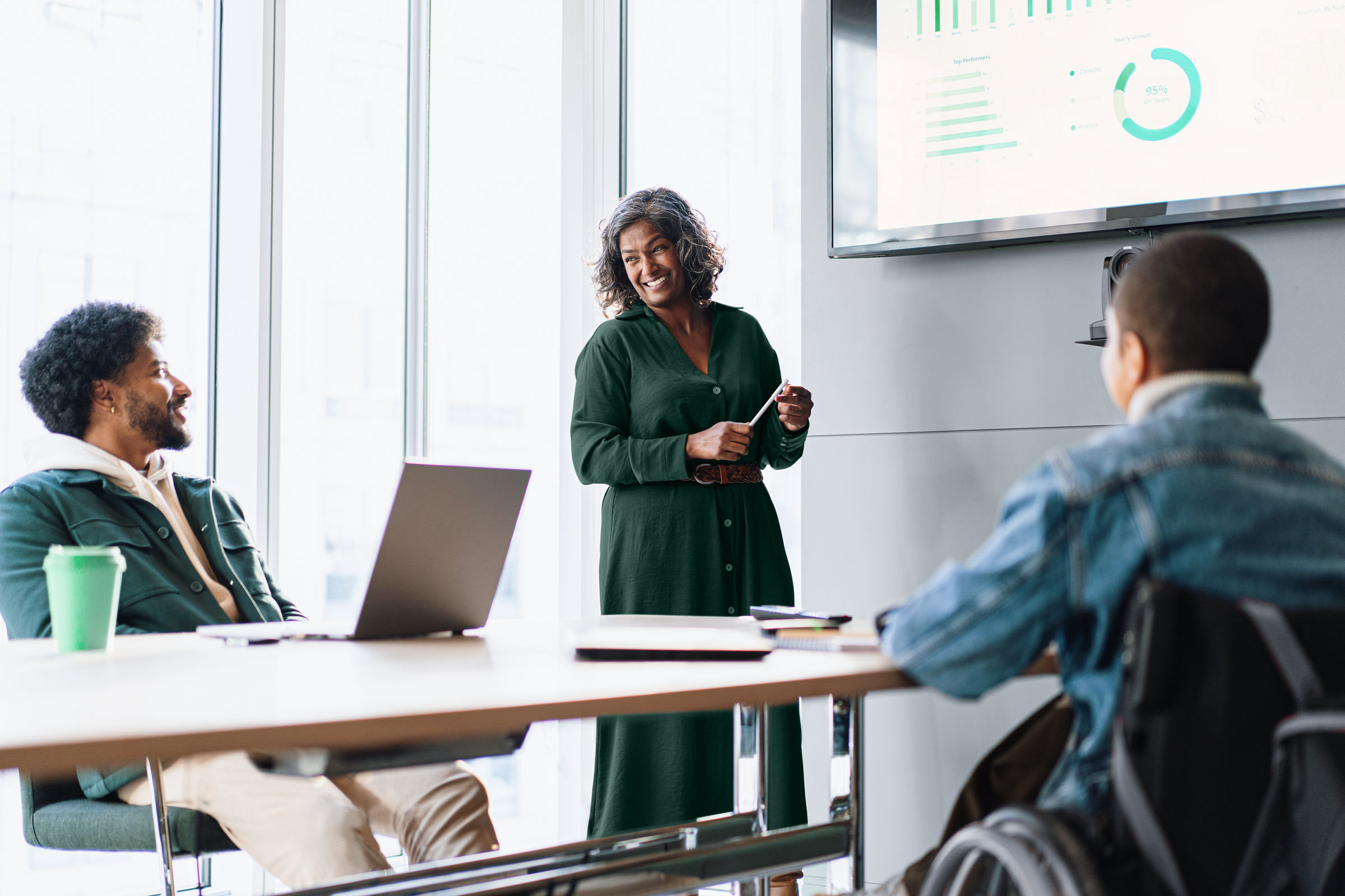 A smiling female presenter is standing at the front of a meeting room near the screen and a smiling male employee and another employee in a wheelchair are seated at a conference table.