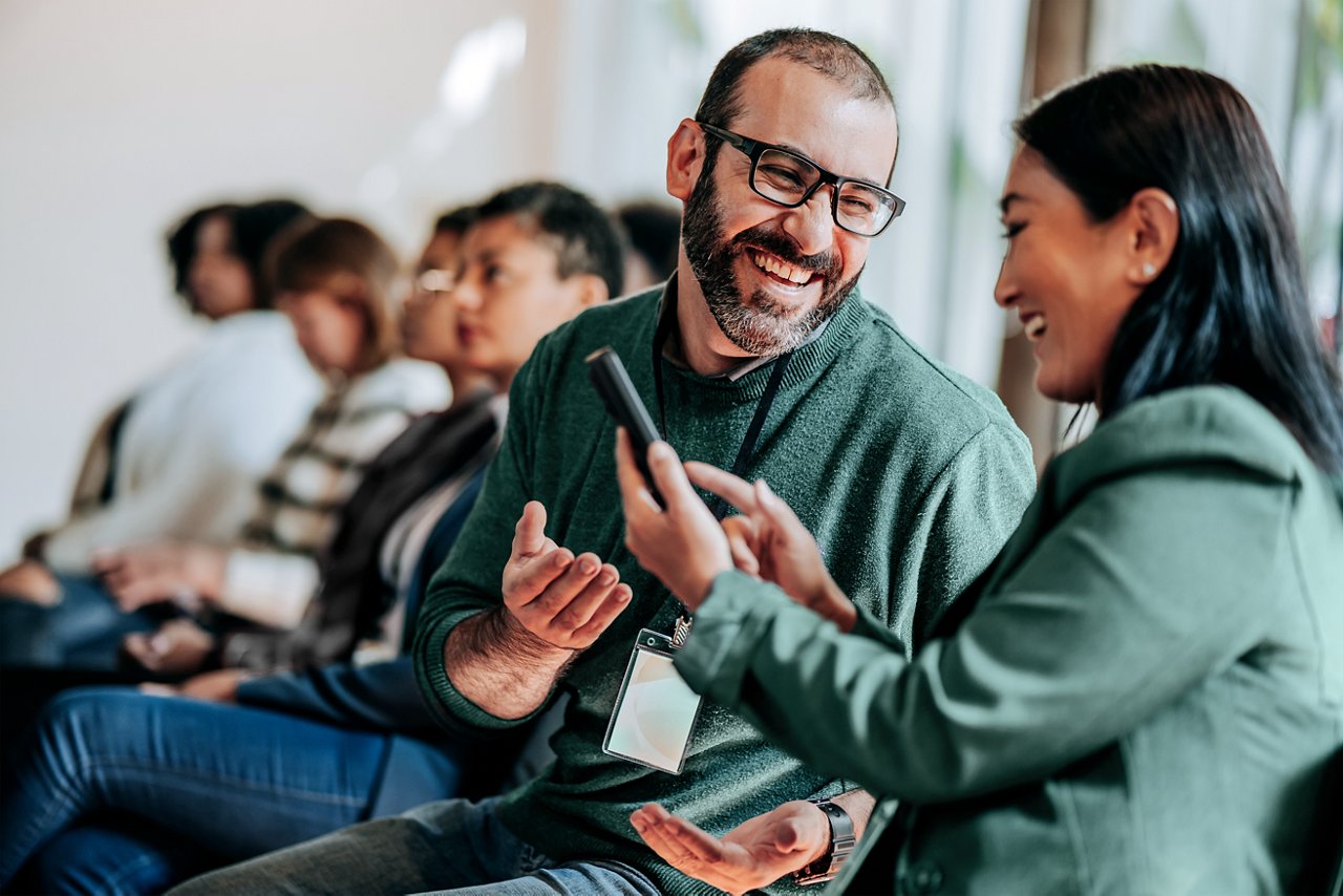 Group of people at a meeting or class with two attendees chatting and viewing a cell phone.