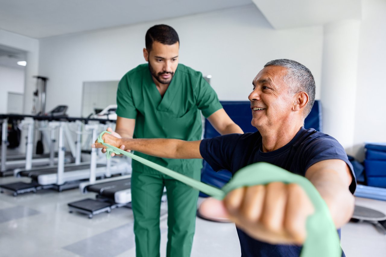 A male physical therapist assisting a male patient during a physical therapy session. The patient is smiling and stretching a band with his arms.