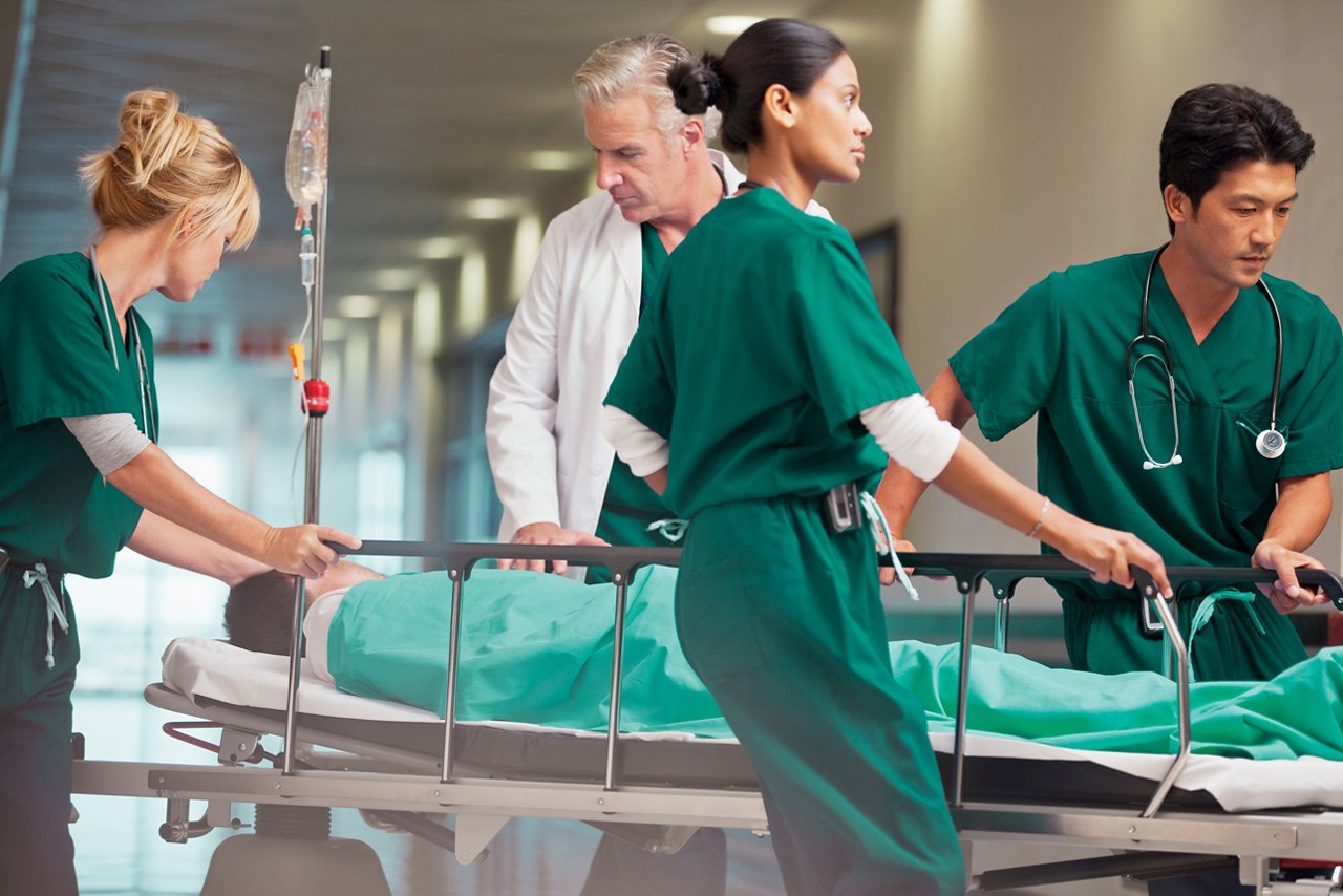 A team of four clinicians rushing a patient on a stretcher down a hospital hallway.