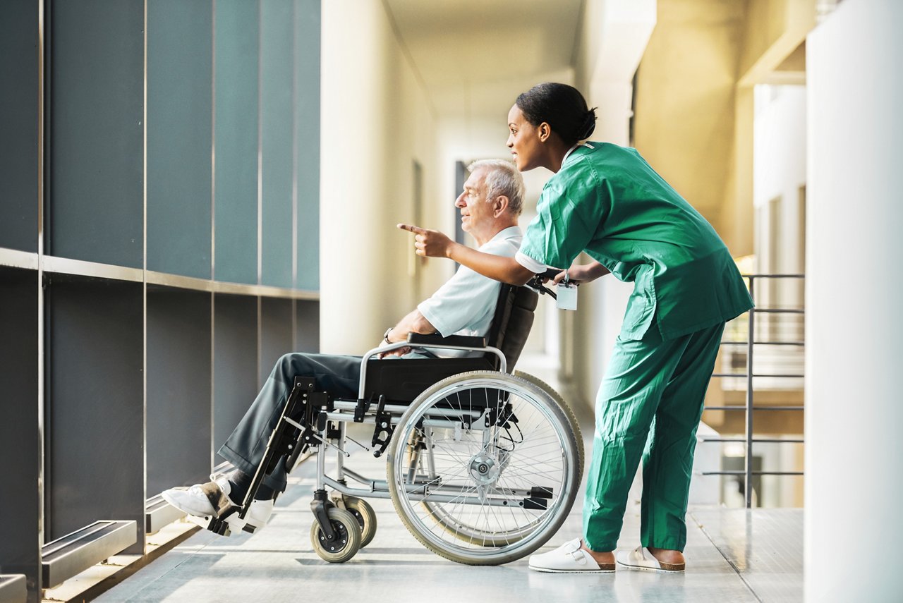 A female clinician standing next to an elderly man in a wheelchair, smiling and pointing at something out a window.