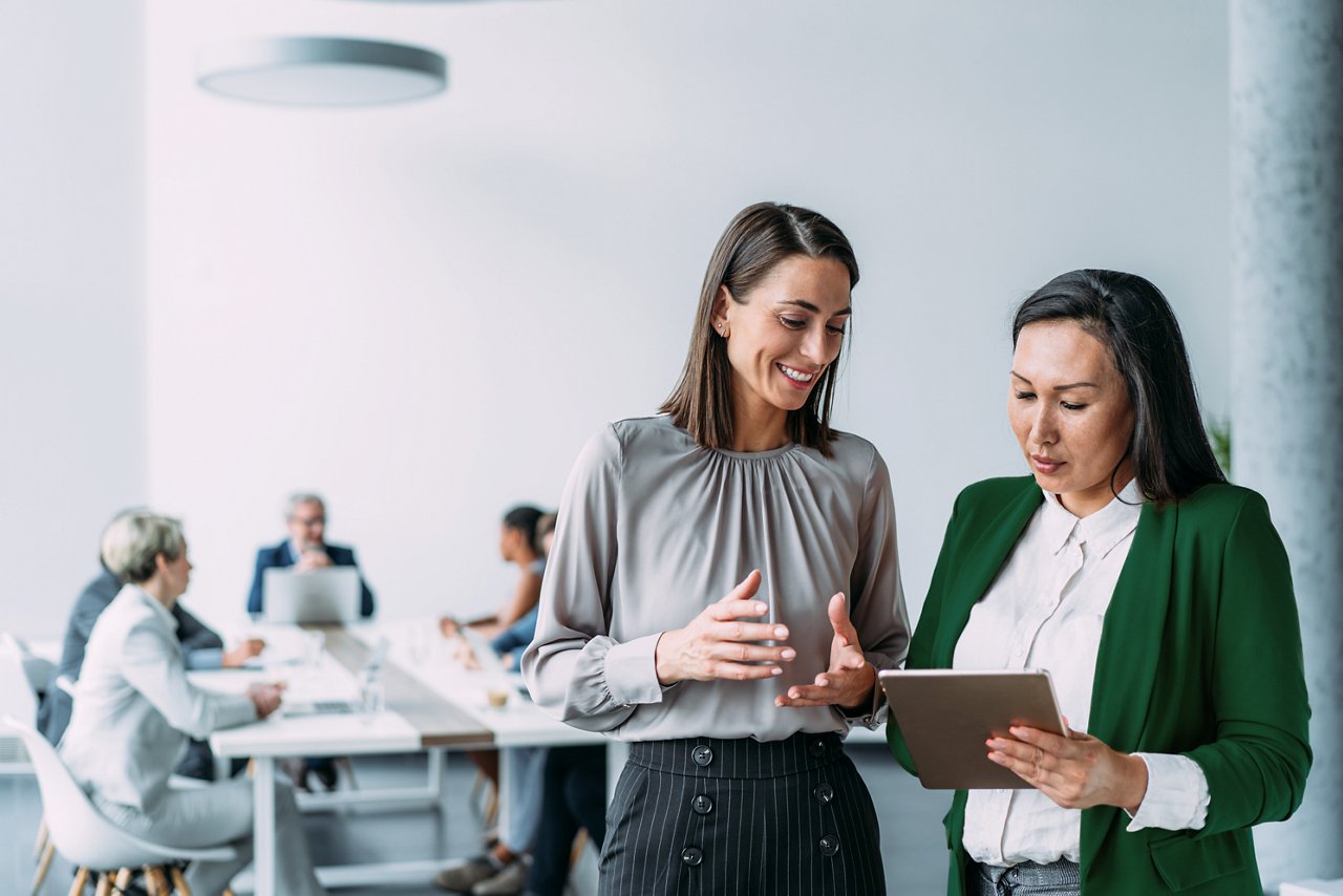 Two woman having a side conversation and viewing a tablet while a group of colleagues are in a meeting.