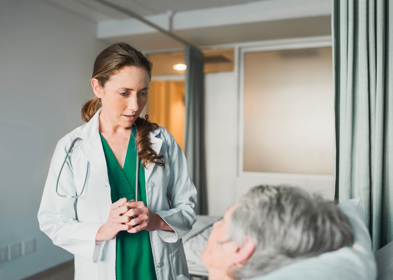 A female clinician explaining something to an elderly patient lying in a hospital bed.