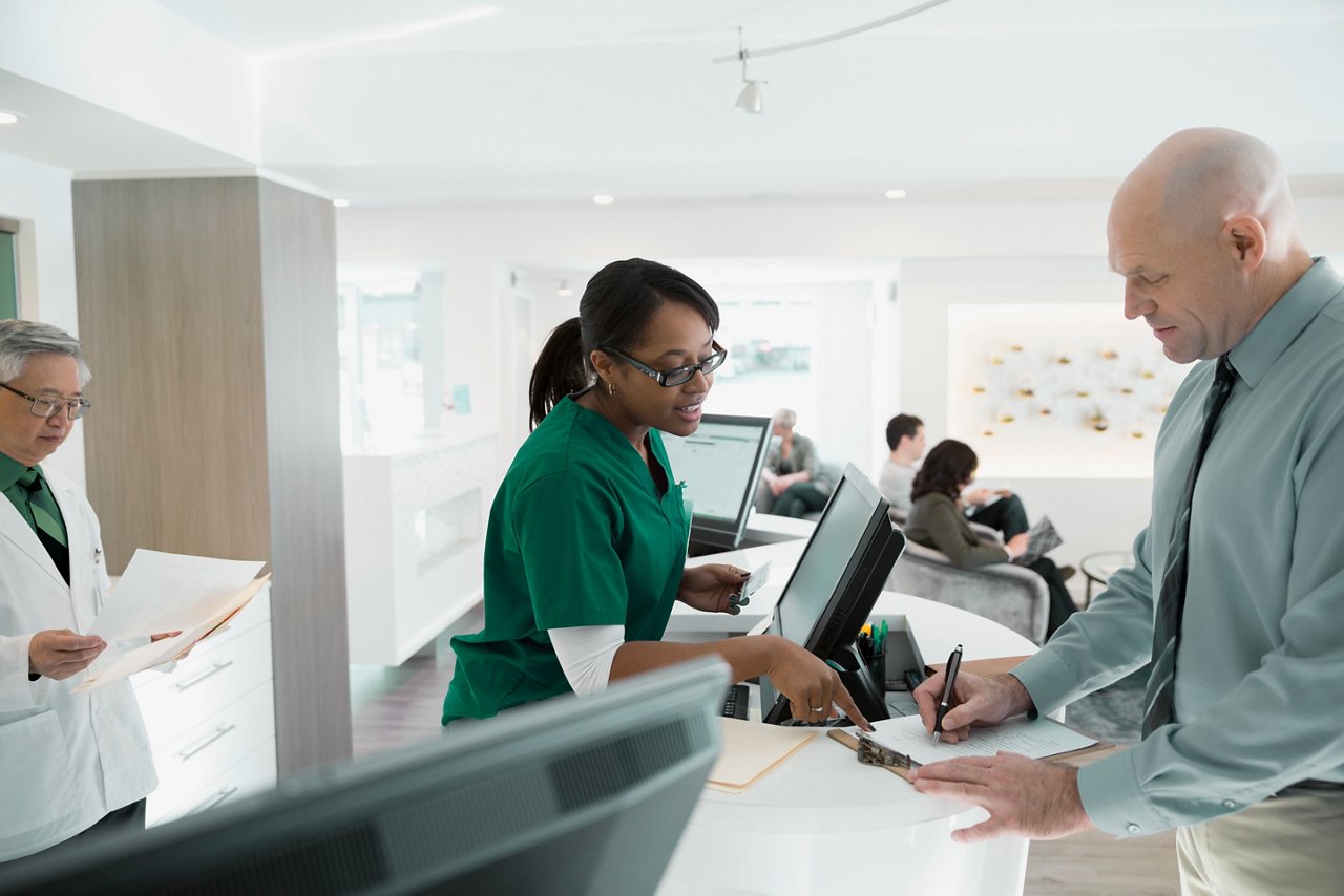 A female working the front desk of a clinician's office helping a male patient fill out forms.