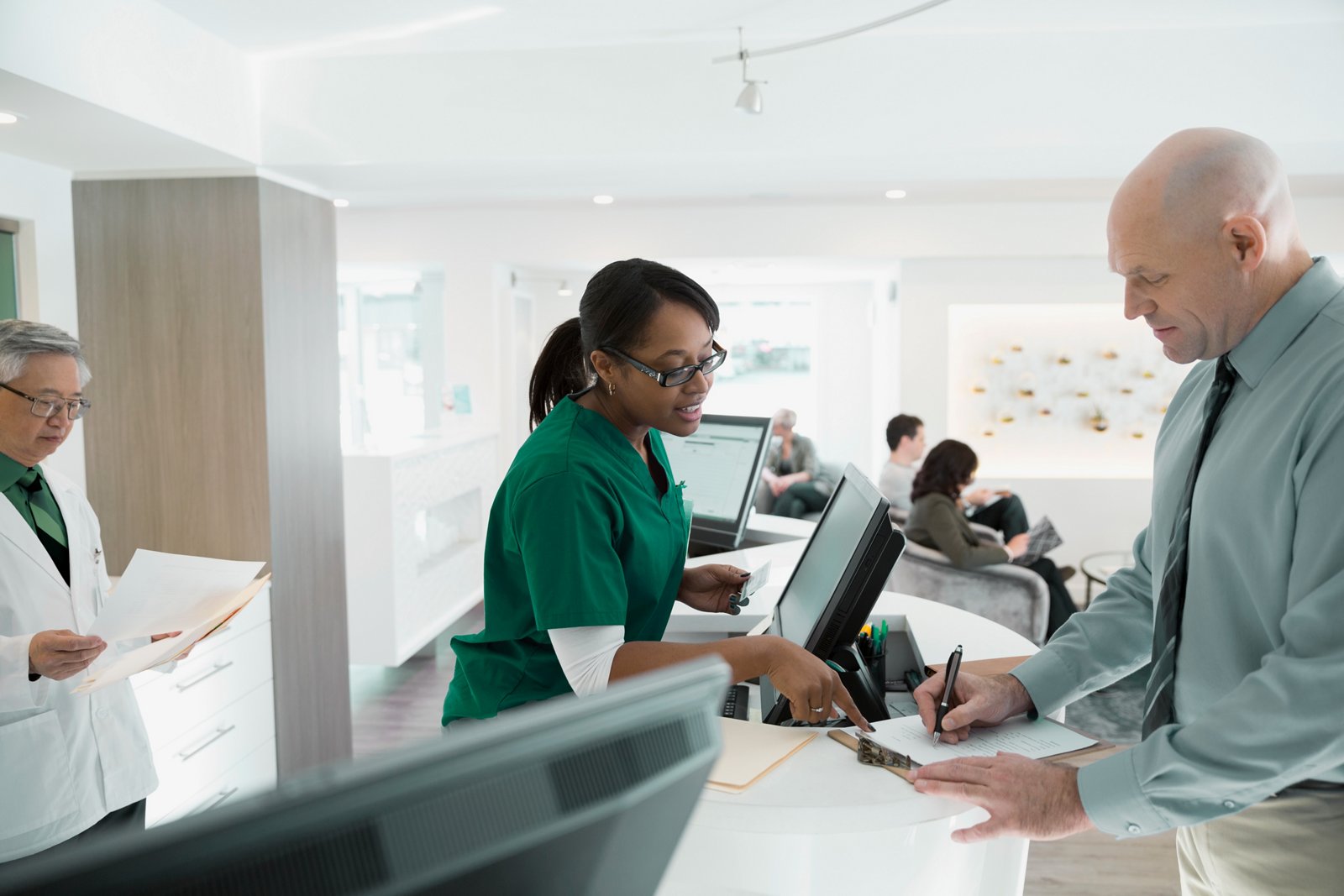 A female working the front desk of a clinician's office helping a male patient fill out forms.