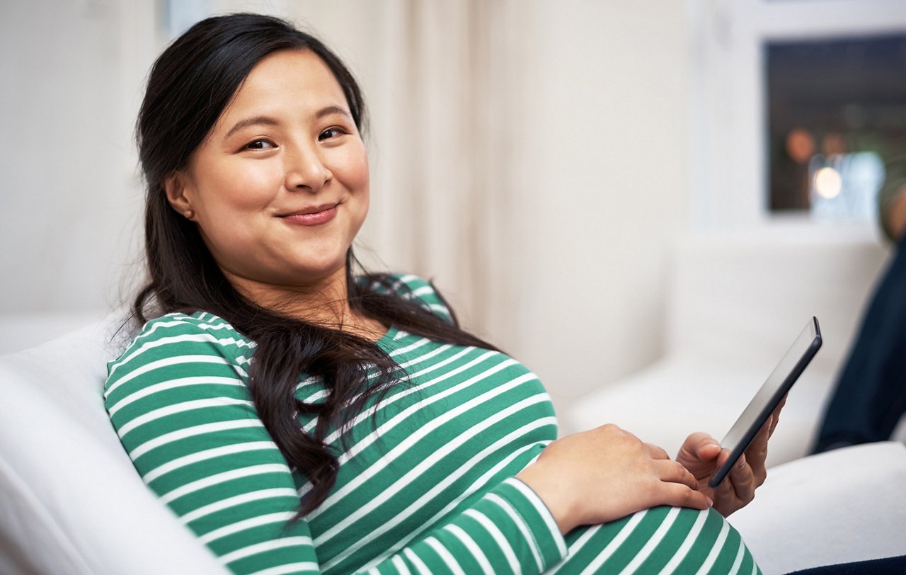 A pregnant woman with her hand on her stomach holding a tablet and smiling.