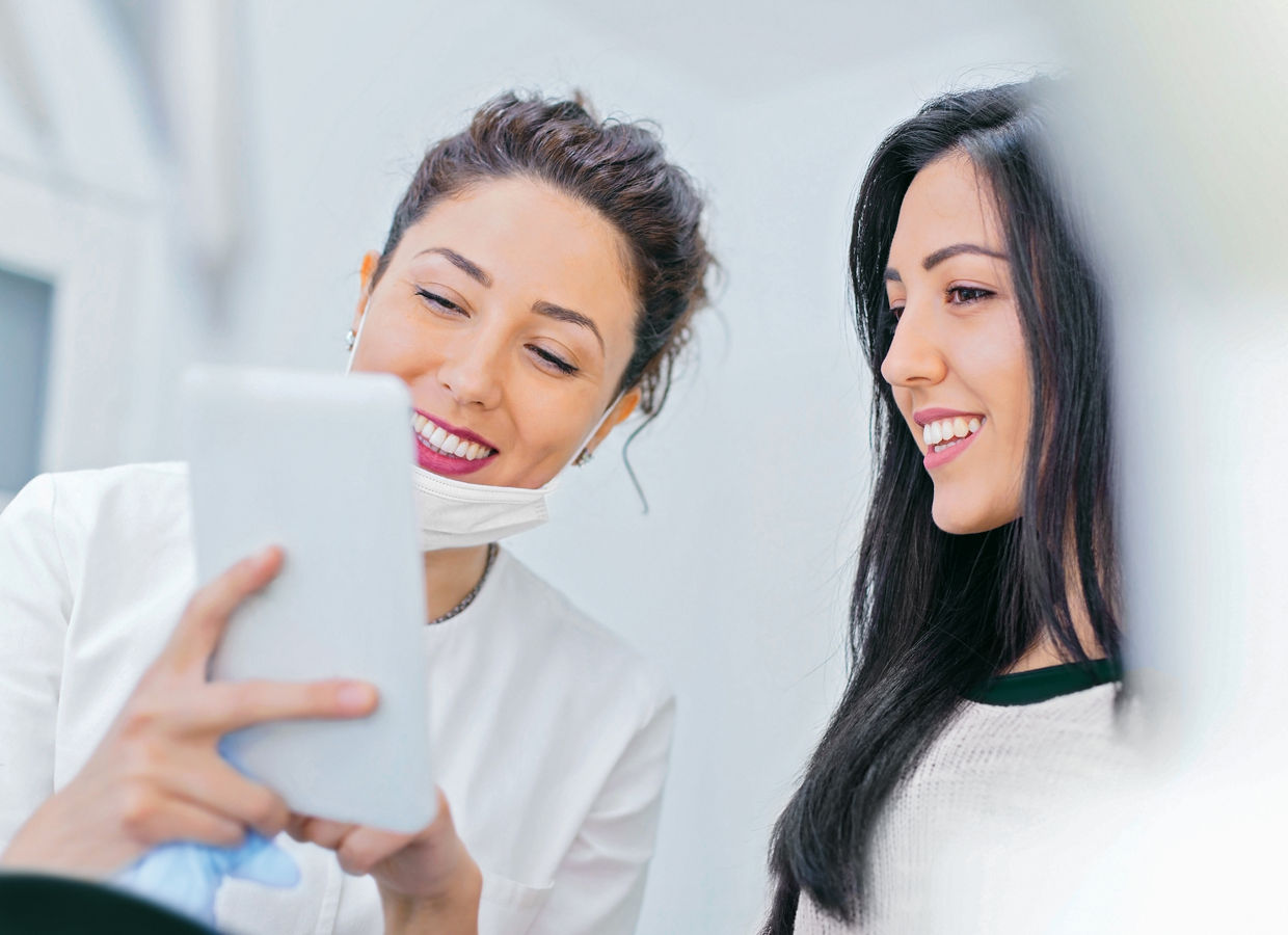 This Getty stock photo has been retouched to reflect the Solventum brand. Photo of a smiling female oral care professional and a smiling female patient interacting while viewing a tablet screen.