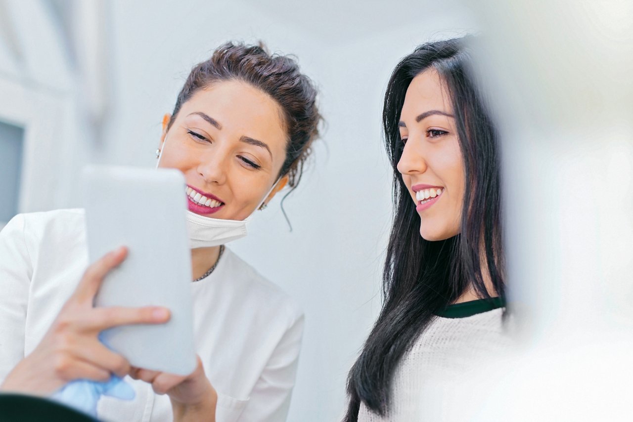 A dentist, with mask pulled down, shows a patient a tablet and points to information on it.