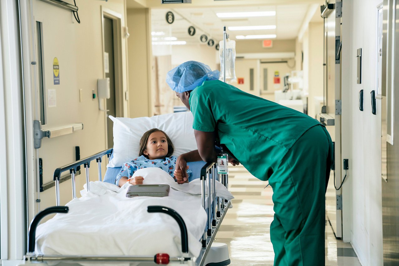 A male clinician in scrubs and a bouffant cap holding the hand of a little girl who is lying in a hospital gurney